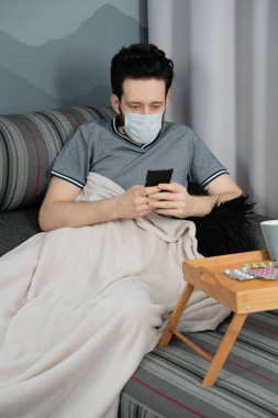 Tıbbi maskeli ve ekoseli bir adam evinde koltukta oturan akıllı telefona bakıyor.