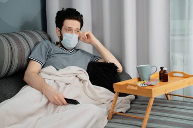 Tıbbi maskeli ve battaniyeli bir adam evde kanepede televizyon seyrediyor. Yakınlarda bir kupa ve ilaç var.