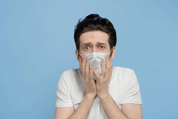 Beyaz tişörtlü ve tıbbi maskeli bir adam korkuyor ve yüzünü mavi arka plana yapıştırıyor.