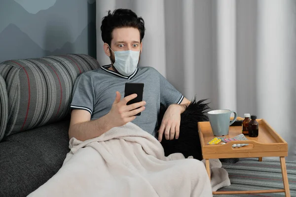 Tıbbi maskeli ve ekoseli bir adam evinde koltukta oturan akıllı telefona bakıyor.
