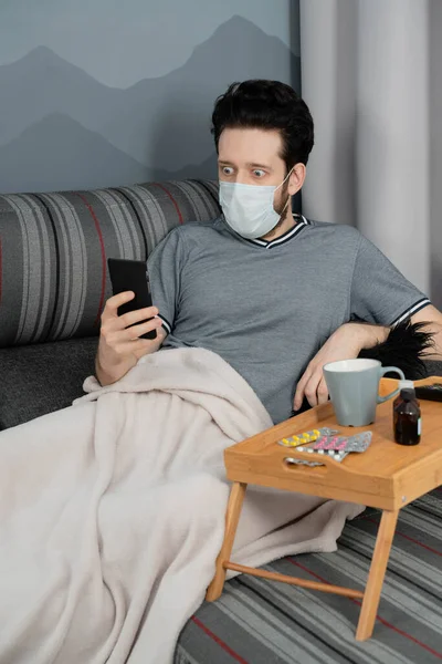 Tıbbi maskeli ve ekoseli bir adam evinde koltukta oturan akıllı telefona bakıyor.