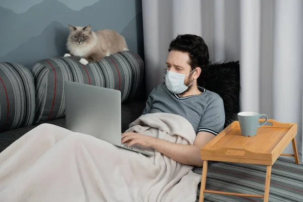 Tıbbi maskeli bir adam evinde, kanepede oturan bir dizüstü bilgisayar üzerinde çalışıyor ve yanında bir kedi oturuyor.