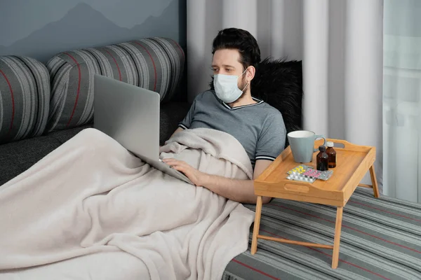 Tıbbi maskeli bir adam evinde, kanepede dizüstü bilgisayarla çalışıyor.