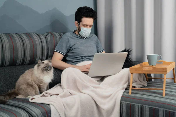 Tıbbi maskeli bir adam evinde, kanepede oturan bir dizüstü bilgisayar üzerinde çalışıyor ve yanında bir kedi oturuyor.