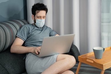 Tıbbi maskeli bir adam evinde, kanepede dizüstü bilgisayarla çalışıyor.