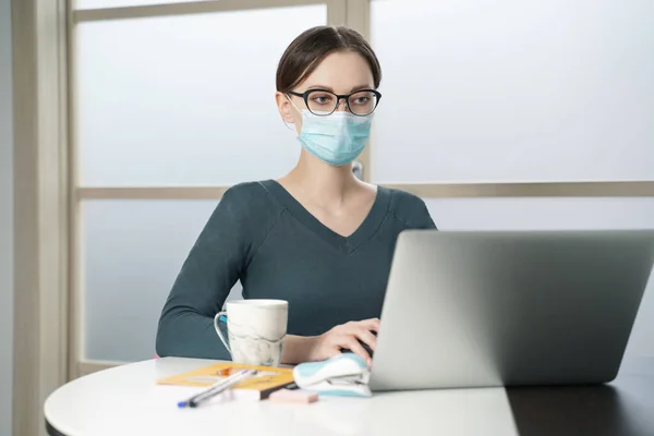 Sağlık maskeli genç bir kadın merkez büroda dizüstü bilgisayarda çalışıyor.