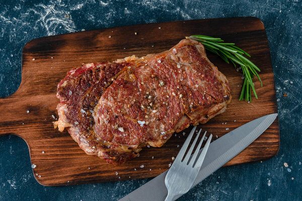 Beef steak with coarse salt and a sprig of rosemary on a wooden cutting board against a dark background