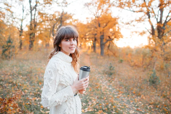 Belle fille dans la forêt d'automne. avec une tasse de boisson chaude. jeunes cheveux longs. Habillé en pull et chapeau — Photo