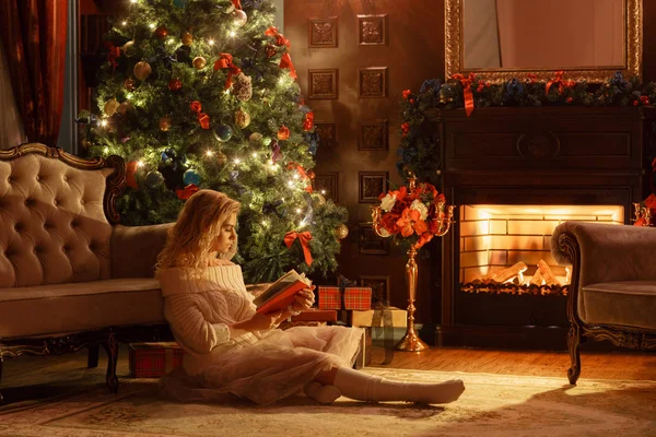 Noite de Natal. Jovem bela mulher loira ler livro em apartamentos clássicos uma lareira, árvore decorada . — Fotografia de Stock