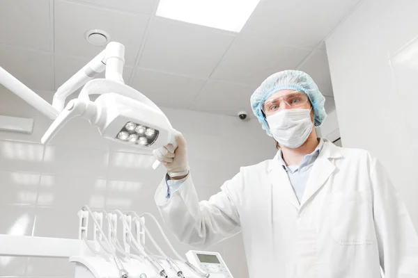 Vista inferior de un médico retratista dentista, cirujano en uniforme y máscara en la cara, con gafas, sosteniendo la lámpara en la mano . — Foto de Stock