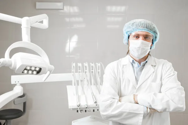 Dentista de pie en el consultorio dental y mirando a la cámara. en la máscara, gafas, un abrigo blanco — Foto de Stock