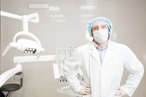 dentist standing in the dental office and looking at camera. in the mask, glasses, a white coat