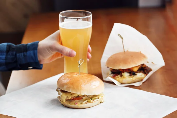 Крупным планом мужчины держат стакан пива в руке. Два восхитительных бургера с луком, помидорами, ломтиком сыра чеддер и салатом с начо-чипсами на доске . — стоковое фото