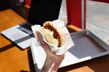 İş yerinde - fast food yeme. Burger dizüstü yakınındaki. çalışırken öğle yemeği molası veya online paylaşım. closeup el holding gıda