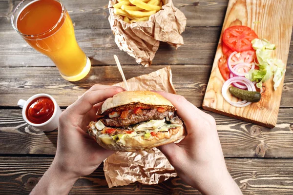 Manos sosteniendo deliciosas hamburguesas frescas con papas fritas, salsa y cerveza en la mesa de madera vista superior . — Foto de Stock