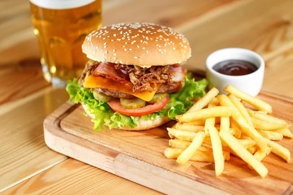 Grande hambúrguer saboroso e batatas fritas com cerveja no fundo na mesa de madeira — Fotografia de Stock