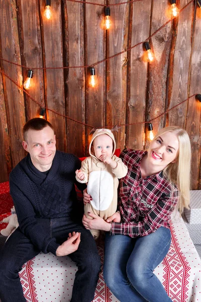 Met Kerstmis en gelukkige familie. De ouders en de baby zit. Muur van houten planken en garland — Stockfoto