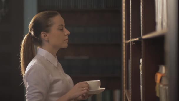 Donne con tazza di caffè. Una studentessa che prende un libro dallo scaffale della biblioteca. mano di piombo sugli scaffali con libri — Video Stock