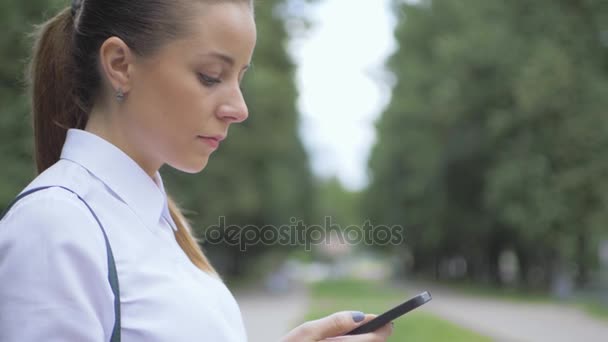 Vista lateral de un teléfono móvil de mensajería de texto de chica universitaria en el parque. primer plano — Vídeo de stock