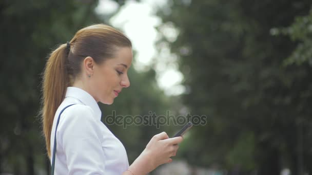 Vista lateral de un teléfono móvil de mensajería de texto de chica universitaria en el parque. primer plano — Vídeo de stock