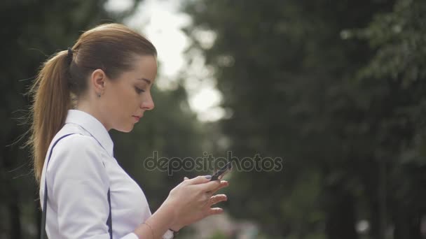 Вид сбоку на девушку из колледжа, переписывающуюся по мобильному телефону в парке. Крупный план — стоковое видео