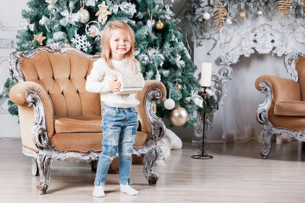 Cute little girl sitting in a chair and opens a box with a present for background Christmas tree with ornaments. — Stock Photo, Image