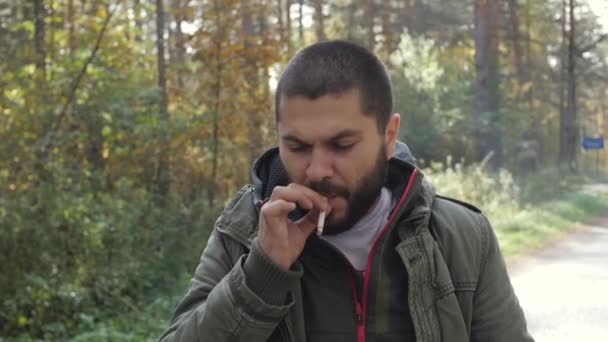 Joven hombre guapo vestido con una chaqueta en un bosque de otoño fuma con confianza un cigarrillo, dejar que el humo . — Vídeos de Stock