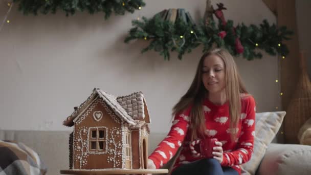 Hermosa mujer con traje de invierno bebiendo té con pan de jengibre en casa cerca del árbol de Navidad — Vídeo de stock