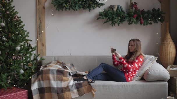 Chica feliz viendo contenido de streaming en línea en un teléfono inteligente sentado en un sofá en invierno en casa — Vídeo de stock