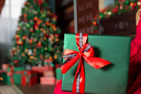 Yeşil Noel hediye yakın çekim ön plan. Kırmızı kurdele yay. Arka plan bulanık ışıkları ve ağaç. kopya alanı — Stok fotoğraf