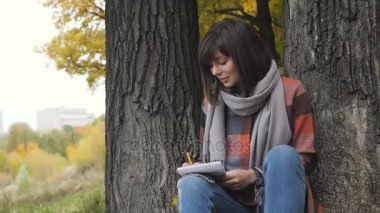 Harika sonbahar görünümü. Büyüleyici genç mutlu bir parkta çimlerde oturan kadın gülümsüyor not not defterinde almak.