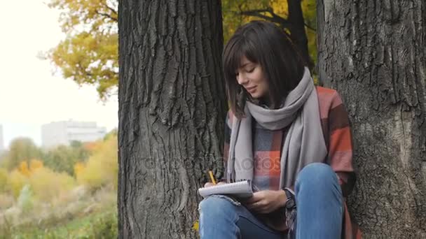 Prachtig herfst uitzicht. Charmante jonge gelukkig lachende vrouw zittend op het gras in een park, het maken van notities in notitieblok. — Stockvideo