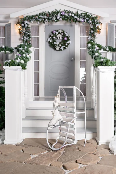 Szánkót és korcsolya az előtérben. ház bejáratának díszített ünnepekre. Karácsonyi dekoráció. Garland fenyő fa ágai és a fények a korlát — Stock Fotó