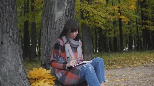 Prachtig herfst uitzicht. Charmante jonge gelukkig lachende vrouw zittend op het gras in een park, het maken van notities in notitieblok. — Stockvideo