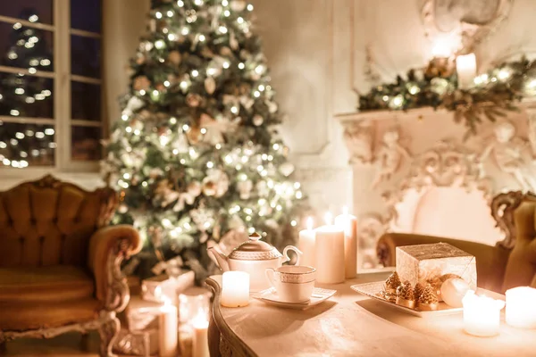 Jul på kvällen med levande ljus. klassiska lägenheter med en vit spis, dekorerade träd, soffa, stora fönster och ljuskrona. — Stockfoto