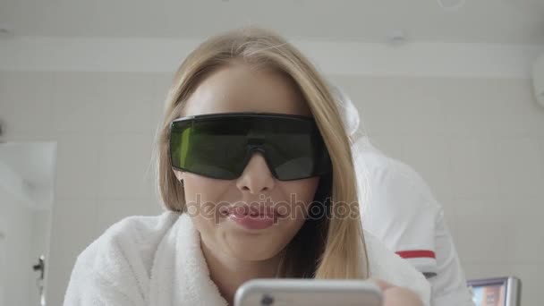 Portrét dívky v brýle, používá telefon. Laserová epilace a kosmetice. Odstranění chloupků na nohách dámy. v kosmetickém beauty clinic spa. — Stock video