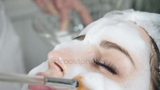 Mujer bonita joven recibiendo tratamientos en salones de belleza. Joven hermosa mujer morena en la oficina esteticista acostado en el sofá. Espuma de limpieza facial usando . — Vídeos de Stock