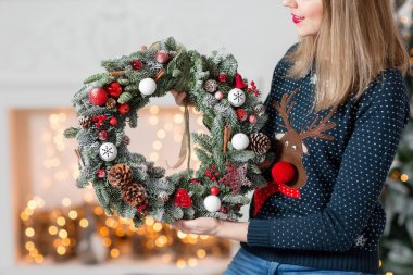 Young woman holding a Christmas wreath with fir branches for the holiday. The new year celebration. on the background of fireplace and Christmas tree, lights, garlands clipart