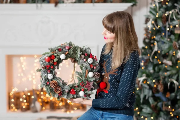 Jonge vrouw met een kroon van Kerstmis met spar takken voor de vakantie. De nieuwe jaarviering. op de achtergrond van de open haard en een kerstboom, lichtjes, slingers — Stockfoto