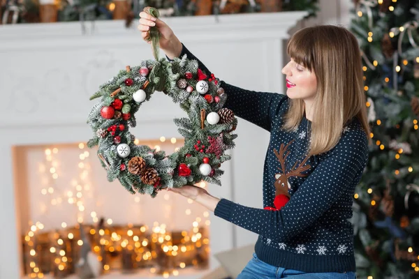 Jonge vrouw met een kroon van Kerstmis met spar takken voor de vakantie. De nieuwe jaarviering. op de achtergrond van de open haard en een kerstboom, lichtjes, slingers — Stockfoto