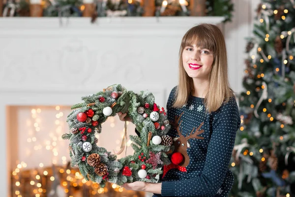 Jonge vrouw met een kroon van Kerstmis met spar takken voor de vakantie. De nieuwe jaarviering. op de achtergrond van de open haard en een kerstboom, lichtjes, slingers — Stockfoto