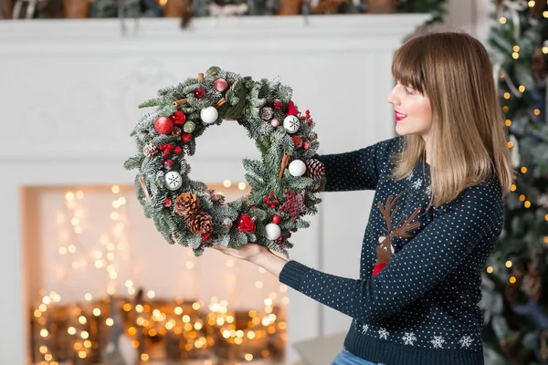 Jonge vrouw met een kroon van Kerstmis met spar takken voor de vakantie. De nieuwe jaarviering. op de achtergrond van de open haard en een kerstboom, lichtjes, slingers — Stockfoto