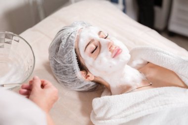 Genç güzel kadın güzellik salonları içinde tedavi alma. Genç güzel esmer kadını kanepede yatan office güzellik uzmanı. Yüz temizleyici köpük kullanarak.
