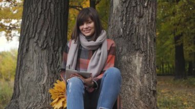 Harika sonbahar görünümü. Sevimli genç mutlu gülümseyen bir parkta çimlerde oturan kadın tablet kullanmak