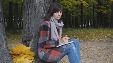 Harika sonbahar görünümü. Büyüleyici genç mutlu bir parkta çimlerde oturan kadın gülümsüyor not not defterinde almak.