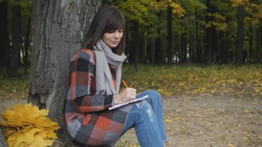 Harika sonbahar görünümü. Büyüleyici genç mutlu bir parkta çimlerde oturan kadın gülümsüyor not not defterinde almak.