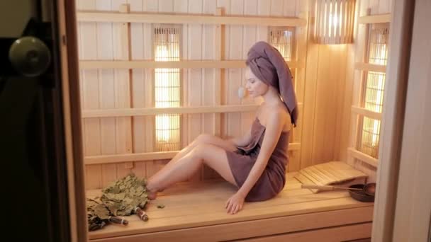 Молодая женщина отдыхает в сауне, одетая в полотенце. Интерьер финской сауны, инфракрасные панели для медицинских процедур, классическая деревянная сауна . — стоковое видео