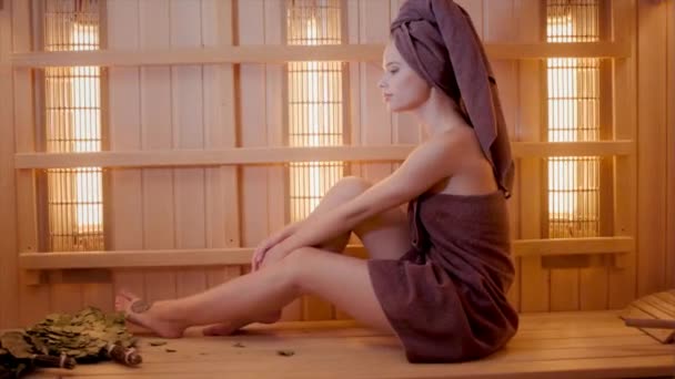 Jonge vrouw ontspannen in een sauna gekleed in een handdoek. Interieur van de nieuwe Finse sauna, infrarood panelen voor medische procedures, klassieke houten sauna. — Stockvideo