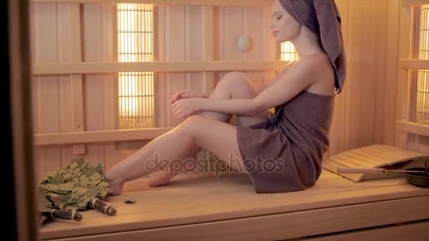 Giovane donna rilassante in una sauna vestita con un asciugamano. Interno della nuova sauna finlandese, pannelli a infrarossi per procedure mediche, classica sauna in legno . — Video Stock
