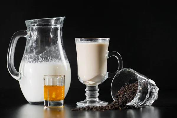 Jest czarna herbata ans mleka z butelki dzbanek do kieliszka na czarnym tle. — Zdjęcie stockowe